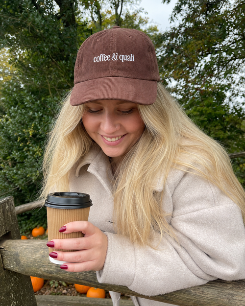 A woman outdoors wearing a brown corduroy cap with 'coffee & quail' embroidered in white, holding a paper coffee cup.