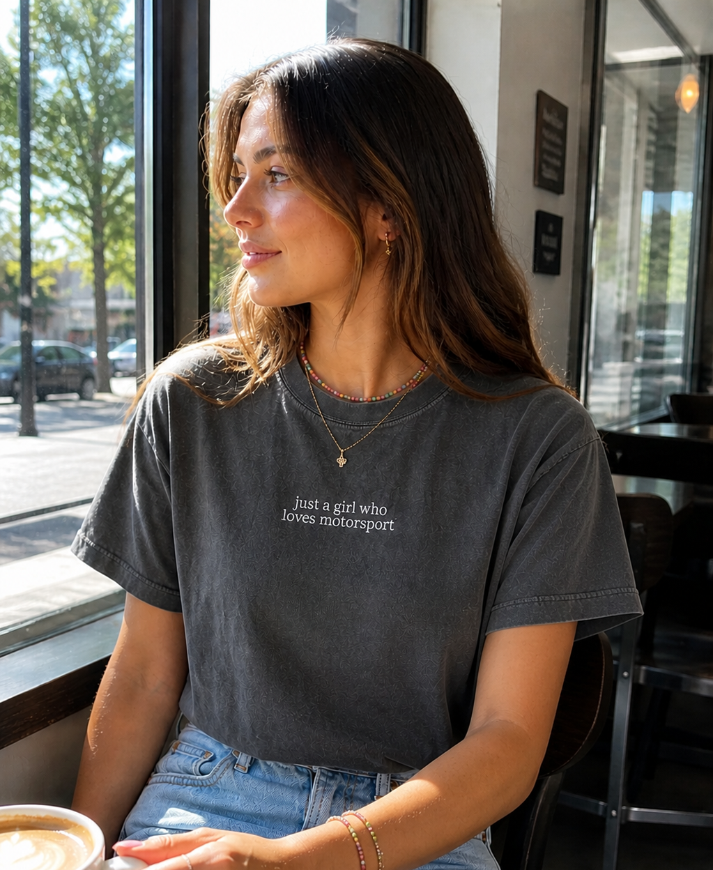 Woman sitting at a cafe table with a coffee, wearing a dark gray t-shirt with center chest embroidery that says "just a girl who loves motorsport"