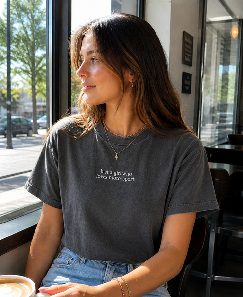 Woman sitting at a cafe table with a coffee, wearing a dark gray t-shirt with center chest embroidery that says "just a girl who loves motorsport"