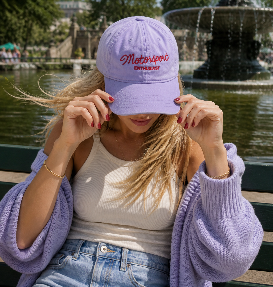 Woman wearing a lavender cap with 'Motorsport Enthusiast' embroidered text, sitting on a bench by a fountain.