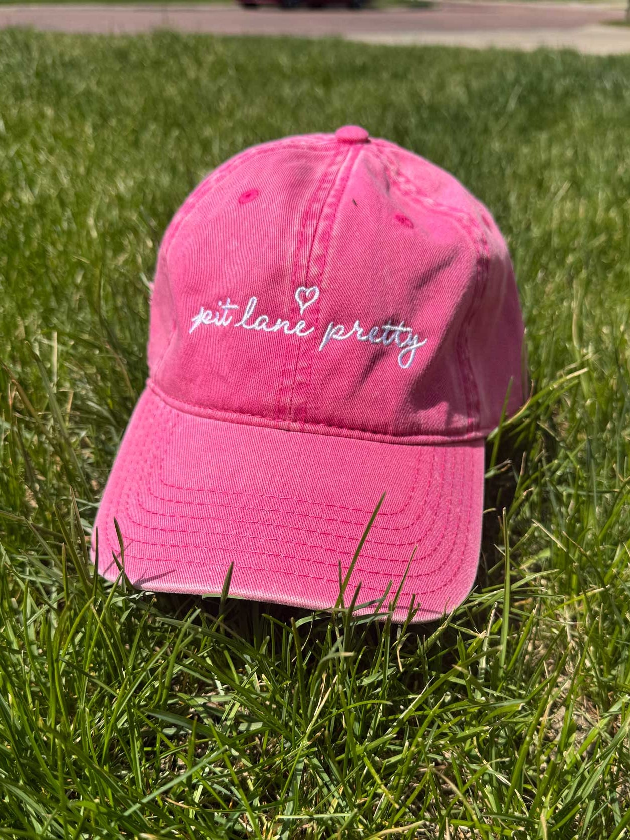 Pink cap with 'pit lane pretty' phrase and heart embroidered text on grass