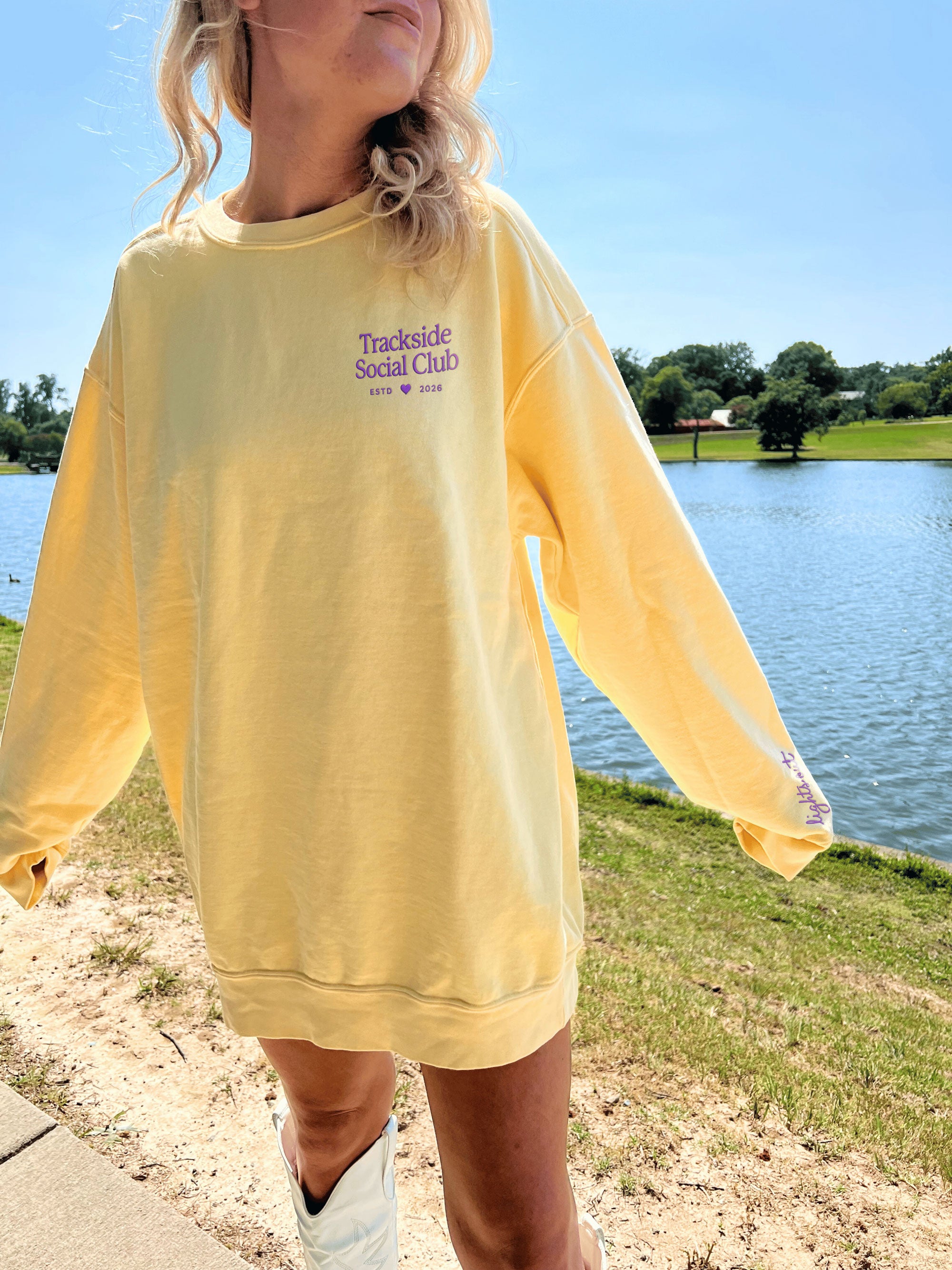 Person wearing a yellow sweatshirt with 'Trackside Social Club' text by a lake.