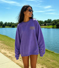 Woman wearing a purple sweatshirt with gold embroidery on left chest that says Trackside Social Club, with embroidery on wrist that says "lights out"